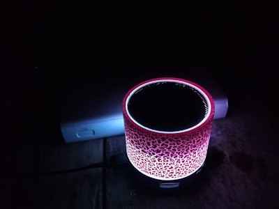 A colorful cylindrical speaker with a textured red and white pattern is illuminated against a dark background. It is placed on a wooden surface next to a partially visible electronic device, possibly a power bank.