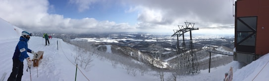A snowy mountain landscape with a wide view of a valley and distant hills. Skiers are standing near a ski lift station, preparing for a descent. The sky is partly cloudy, casting shadows on the snow-covered terrain.