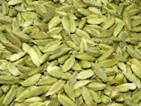 A large quantity of green cardamom pods are spread out together, displaying their smooth, ribbed texture. The pods vary slightly in shade, ranging from light to dark green, and have a natural, organic appearance.