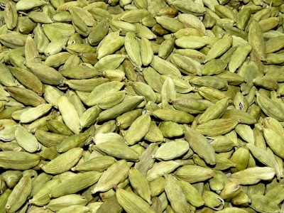 A large quantity of green cardamom pods are spread out together, displaying their smooth, ribbed texture. The pods vary slightly in shade, ranging from light to dark green, and have a natural, organic appearance.