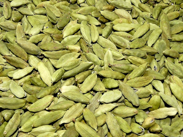 A large quantity of green cardamom pods are spread out together, displaying their smooth, ribbed texture. The pods vary slightly in shade, ranging from light to dark green, and have a natural, organic appearance.