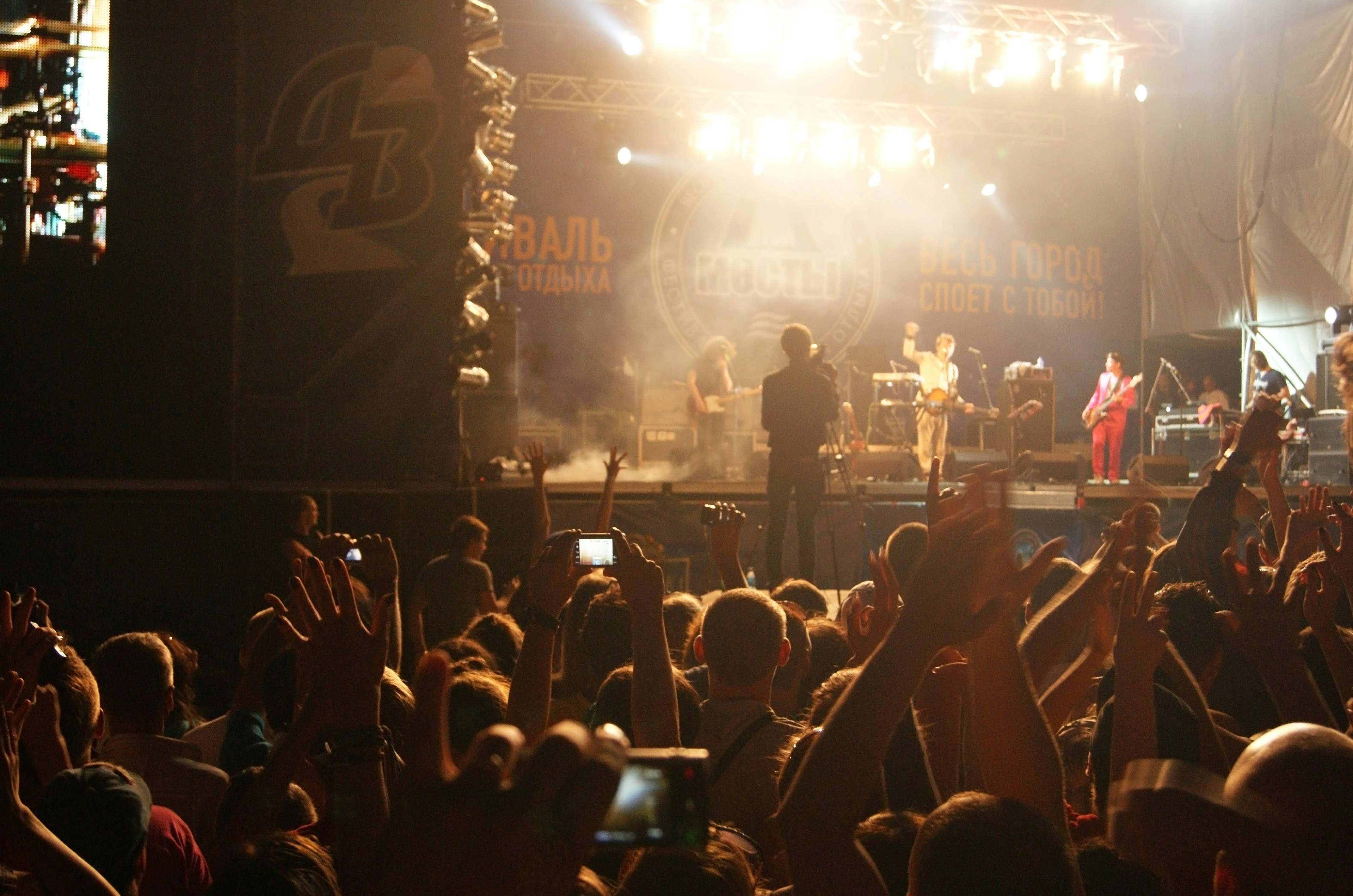 Concert audience with hands raised, bright stage lights above, energetic crowd atmosphere