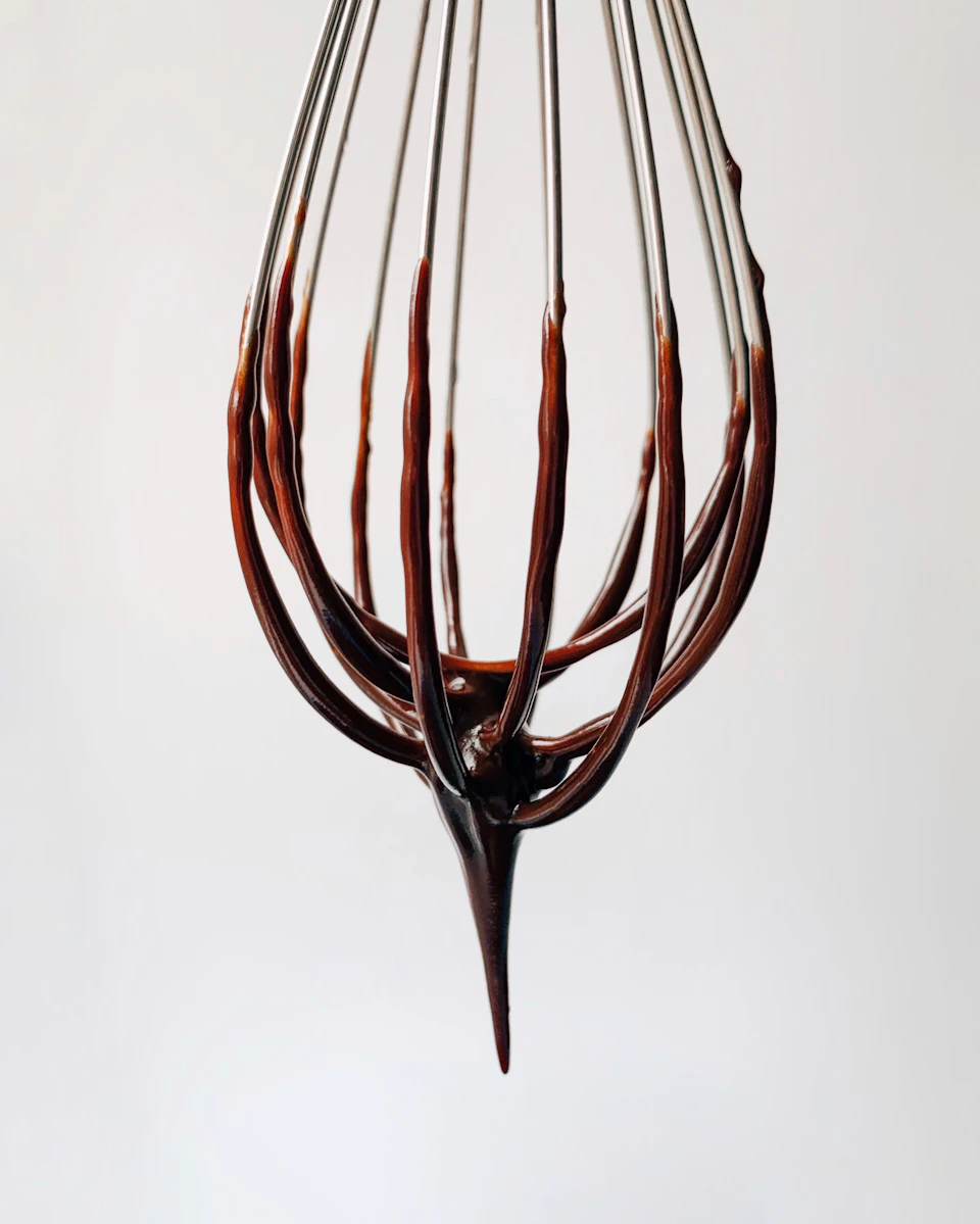 The image shows a close-up view of a chocolate drizzled wire whisk against a white background. The chocolate is brown and appears to be dripping from the whisk, creating an artistic effect on the wire.