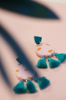 A close-up of colorful earrings with a playful design, featuring circular pink shapes with yellow spots and teal tassels. The earrings are partially obscured by blurred green leaves in the foreground, adding a touch of nature to the scene.