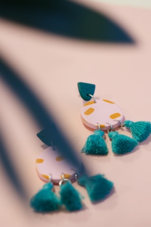 A close-up of colorful earrings with a playful design, featuring circular pink shapes with yellow spots and teal tassels. The earrings are partially obscured by blurred green leaves in the foreground, adding a touch of nature to the scene.