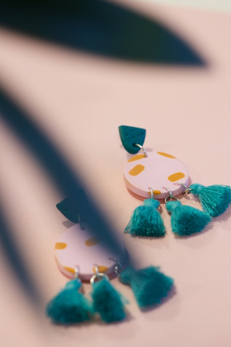 A close-up of colorful earrings with a playful design, featuring circular pink shapes with yellow spots and teal tassels. The earrings are partially obscured by blurred green leaves in the foreground, adding a touch of nature to the scene.