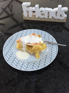 A slice of pie with cream on a decorative plate, accompanied by a fork. In the background, there is a decorative sign spelling the word 'Friends' with a message below it.