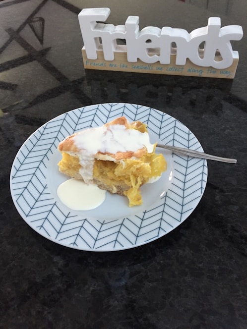 A slice of pie with cream on a decorative plate, accompanied by a fork. In the background, there is a decorative sign spelling the word 'Friends' with a message below it.