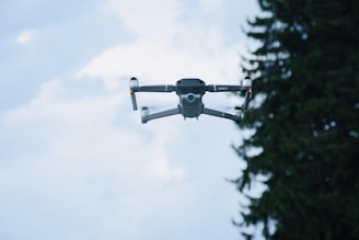 A drone is captured mid-flight against a bright sky, with a few clouds visible in the background. The drone's design is sleek and modern, showcasing its four propellers and compact body. To the right, the silhouette of a pine tree is partially visible.