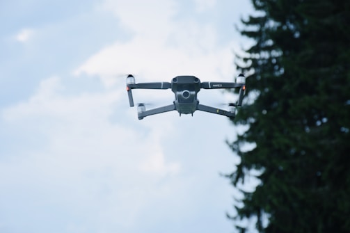 A drone is captured mid-flight against a bright sky, with a few clouds visible in the background. The drone's design is sleek and modern, showcasing its four propellers and compact body. To the right, the silhouette of a pine tree is partially visible.
