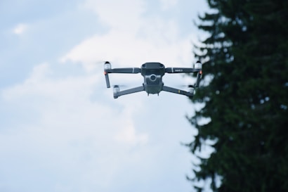 A drone is captured mid-flight against a bright sky, with a few clouds visible in the background. The drone's design is sleek and modern, showcasing its four propellers and compact body. To the right, the silhouette of a pine tree is partially visible.