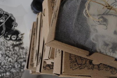 A stack of brown cardboard boxes with intricate black designs on their sides is placed on a surface. A large sheet with detailed black and white drawings is visible to the side. The boxes appear to be packaging or delivery items featuring creative illustrations.