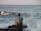 A man in a navy sweatshirt sitting on a rocky beach with waves crashing behind him.