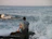 A man in a navy sweatshirt sitting on a rocky beach with waves crashing behind him.