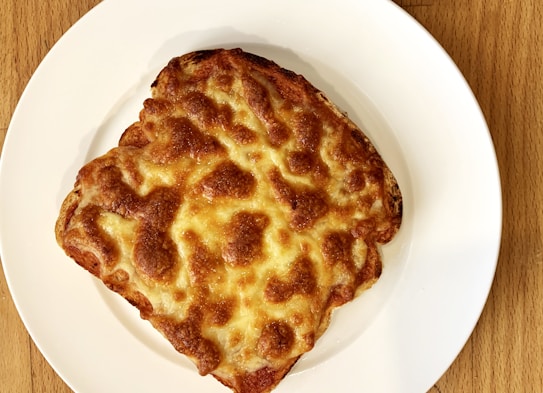 A toasted, golden-brown sandwich or piece of bread topped with melted cheese, arranged on a plain white plate sitting on a wooden surface.