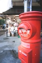 A traditional red Japanese post box is prominently displayed in the foreground, with Japanese writing on it. In the background, there is a street scene with a shop visible, featuring various goods and items outside.