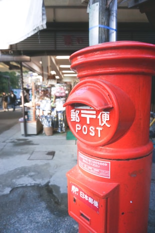 A traditional red Japanese post box is prominently displayed in the foreground, with Japanese writing on it. In the background, there is a street scene with a shop visible, featuring various goods and items outside.