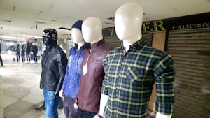 Several mannequins are dressed in different casual outfits, displayed in a row inside a retail setting. The mannequins are wearing a variety of clothing items including plaid shirts, leather jackets, and a beanie hat. The background features other mannequins and a partially closed store with a sign.