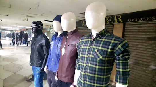 Several mannequins are dressed in different casual outfits, displayed in a row inside a retail setting. The mannequins are wearing a variety of clothing items including plaid shirts, leather jackets, and a beanie hat. The background features other mannequins and a partially closed store with a sign.