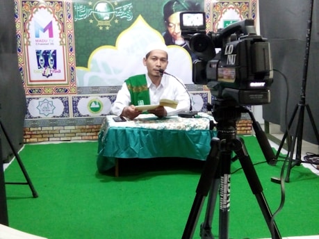 A man wearing a white shirt and a green sash is sitting at a desk covered with a patterned green tablecloth. He is speaking into a microphone and holding a book. In front of him is a large camera on a tripod. The background features decorative patterns and logos.