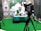 A man wearing a white shirt and a green sash is sitting at a desk covered with a patterned green tablecloth. He is speaking into a microphone and holding a book. In front of him is a large camera on a tripod. The background features decorative patterns and logos.
