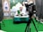 A man wearing a white shirt and a green sash is sitting at a desk covered with a patterned green tablecloth. He is speaking into a microphone and holding a book. In front of him is a large camera on a tripod. The background features decorative patterns and logos.