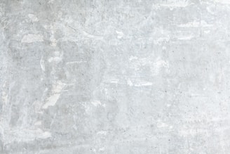 A textured concrete wall with a light gray, slightly weathered surface showing subtle variations and irregularities. The surface exhibits patches of lighter and darker gray, portraying a rustic and industrial look.