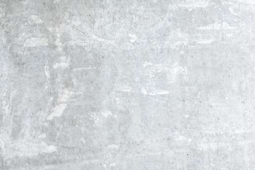 A textured concrete wall with a light gray, slightly weathered surface showing subtle variations and irregularities. The surface exhibits patches of lighter and darker gray, portraying a rustic and industrial look.