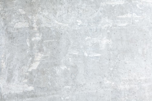A textured concrete wall with a light gray, slightly weathered surface showing subtle variations and irregularities. The surface exhibits patches of lighter and darker gray, portraying a rustic and industrial look.