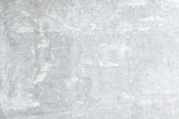 A textured concrete wall with a light gray, slightly weathered surface showing subtle variations and irregularities. The surface exhibits patches of lighter and darker gray, portraying a rustic and industrial look.