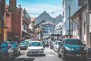 A busy street lined with parked cars and a few vehicles moving along. On the left, there are signs for a hotel and other businesses. The background features a distinct white building with domed architecture, resembling a mosque. The sky is clear and blue, contributing to a warm, sunny atmosphere.