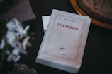 A book titled 'Le Lambeau' by Philippe Lançon rests on a dark surface. The cover is predominantly white with red and black text. The book appears slightly worn, suggesting it has been read or handled. Surrounding the book are blurred objects, including what looks like a plant and part of a decorative item, adding to the intimate and cozy atmosphere of the scene.
