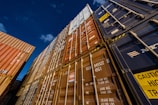 Several stacked shipping containers of various colors including blue, brown, and orange are seen from a low angle against a clear night sky. The containers are marked with different codes and logos, suggesting a busy industrial shipping environment.