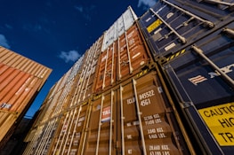 Several stacked shipping containers of various colors including blue, brown, and orange are seen from a low angle against a clear night sky. The containers are marked with different codes and logos, suggesting a busy industrial shipping environment.