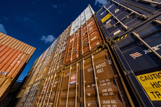 Several stacked shipping containers of various colors including blue, brown, and orange are seen from a low angle against a clear night sky. The containers are marked with different codes and logos, suggesting a busy industrial shipping environment.