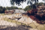 A fenced area contains a pile of rusty and weathered metal scrap pieces, including large beams and angular structures. The background shows lush green trees and grass, with a clear blue sky above.