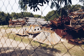 A fenced area contains a pile of rusty and weathered metal scrap pieces, including large beams and angular structures. The background shows lush green trees and grass, with a clear blue sky above.