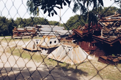 A fenced area contains a pile of rusty and weathered metal scrap pieces, including large beams and angular structures. The background shows lush green trees and grass, with a clear blue sky above.