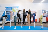 Several colorful prosthetic legs with intricate designs are displayed on stands. In the background, three people are intently observing an exhibition with informational displays about accessibility and assistive technology. A wheelchair is visible to the side.