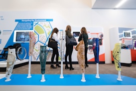 Several colorful prosthetic legs with intricate designs are displayed on stands. In the background, three people are intently observing an exhibition with informational displays about accessibility and assistive technology. A wheelchair is visible to the side.