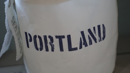 A white object with the word 'PORTLAND' printed in bold, navy blue, slightly distressed letters. To the left, a partially visible tag with more text is attached.