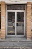 A glass double door entrance with metal frames and handles is set within a brick facade. The reflective glass shows a faint reflection of the street with a parked car and some trees. The ground in front of the door is concrete, with some grass and debris visible around the edges.