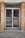 A glass double door entrance with metal frames and handles is set within a brick facade. The reflective glass shows a faint reflection of the street with a parked car and some trees. The ground in front of the door is concrete, with some grass and debris visible around the edges.