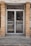 A glass double door entrance with metal frames and handles is set within a brick facade. The reflective glass shows a faint reflection of the street with a parked car and some trees. The ground in front of the door is concrete, with some grass and debris visible around the edges.