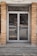 A glass double door entrance with metal frames and handles is set within a brick facade. The reflective glass shows a faint reflection of the street with a parked car and some trees. The ground in front of the door is concrete, with some grass and debris visible around the edges.