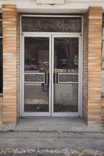 A glass double door entrance with metal frames and handles is set within a brick facade. The reflective glass shows a faint reflection of the street with a parked car and some trees. The ground in front of the door is concrete, with some grass and debris visible around the edges.