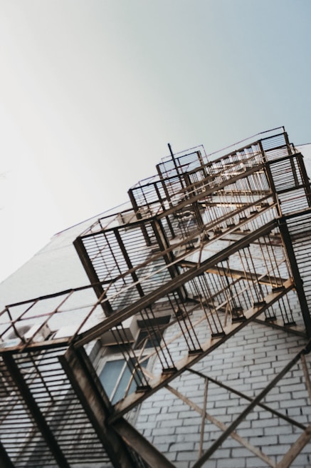 A perspective view of an outdoor metal fire escape attached to the side of a brick building, reaching upwards towards the sky. The ladder is set against a backdrop of light blue sky and the bricks exhibit a pale, worn texture. The structure casts intricate shadows on the building surface, highlighting the grid-like pattern of the metal.