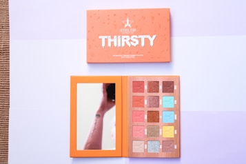 A cosmetics palette with an open lid displaying a mirror on the left and an array of colorful eyeshadow shades on the right. The palette has a bright orange exterior and muted pastel shades inside. Various rectangular and square eyeshadow pans are visible in colors such as brown, blue, pink, and gold. Above the open palette, a closed lid is visible with the brand name and the word 'Thirsty' printed in bold white letters against an orange background.