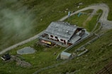 A rustic building with a wooden roof is nestled within a grassy landscape. It is surrounded by a fence and situated near a gravel path that curves alongside the property. There are small animal enclosures visible and a few animals grazing nearby. Mist lightly covers parts of the green hillside, adding to the serene atmosphere.