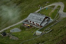 A rustic building with a wooden roof is nestled within a grassy landscape. It is surrounded by a fence and situated near a gravel path that curves alongside the property. There are small animal enclosures visible and a few animals grazing nearby. Mist lightly covers parts of the green hillside, adding to the serene atmosphere.